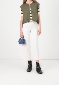 Olivgrüne ärmellose Jacke, gestreiftes gelb-braunes Oberteil, weiße Hose mit weitem Bein, schwarze Stiefeletten mit silbernen Details und blaue Mini-Tasche.