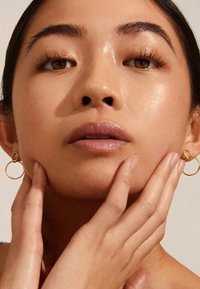 Detailní záběr na ženu s hladkou pletí, zlatými kroužkovými náušnicemi a prsty jemně dotýkajícími se její čelisti a krku na neutrálním pozadí.