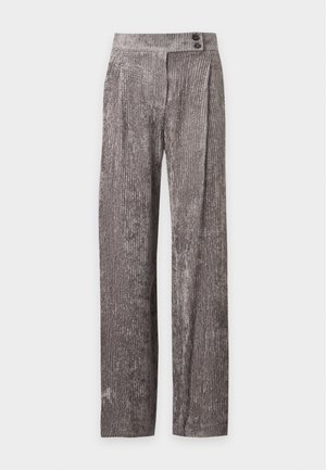 Femme portant un haut beige sans manches et un pantalon ample gris, tenant un sac à main beige, debout devant un fond uni clair.