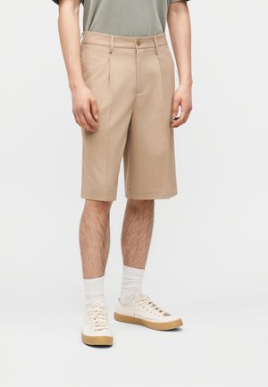 Muž nosi béžové na mieru šité šortky po kolená, biele ponožky a biele nízke tenisky na bielom pozadí.