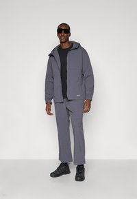 Giacca grigia con cappuccio e tasche frontali, abbinata a pantaloni grigi coordinati. Camicia nera sotto e calzature nere. Finitura elegante e design minimale.