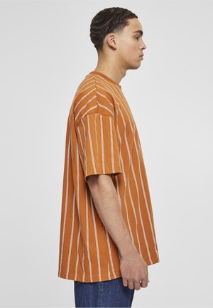 Ung mann i profil som har på seg en oransje kortermet T-skjorte med vertikale hvite striper og blå jeans mot en ensfarget bakgrunn.