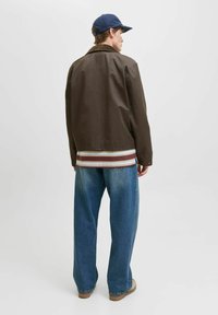 Giacca marrone con colletto in pelliccia, indossata sopra una camicia a righe e abbinata a jeans blu larghi. Presenta un berretto blu navy semplice e scarpe color cammello.