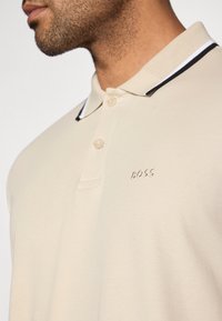 Beige polo tričko s límečkem, který je zakončen černobílým pruhem. Vyšitá značka "BOSS" na levém prsu. Měkký, strukturovaný materiál.