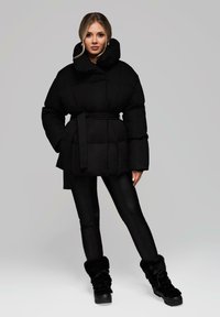 Černá péřová bunda s širokým límcem, opásaným pasem a prošívanou texturou. V kombinaci s černými legínami a černými botami s kožíškem.