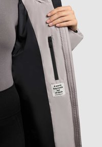 Giacca esterna grigio chiaro con interno nero. Presenta una tasca con zip e un'etichetta che recita: "Un caldo abbraccio fatto di stoffa."