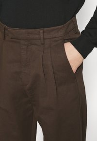 Närbild av en person i bruna veckade byxor, med en hand i fickan, kompletterad av en svart långärmad topp.