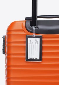 Maleta rígida naranja con superficie texturizada, mango retráctil y una etiqueta negra de equipaje que muestra campos para la información de contacto.