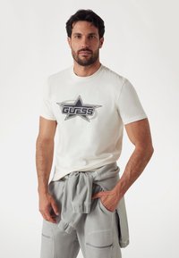 Fehér pamut póló, nagy fekete és szürke csillag grafikával, középen "GUESS" felirattal; szürke nadrággal párosítva, az egyik kezében a zsebében.