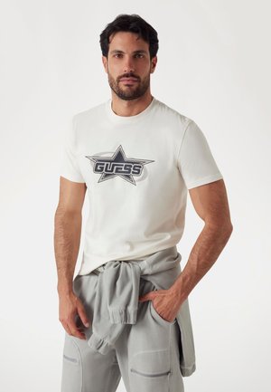 Bijela pamučna majica s velikim grafičkim uzorkom zvijezde u crnoj i sivoj boji sa tekstom "GUESS" u središtu; uparen s sivim hlačama, jednom rukom u džepu.