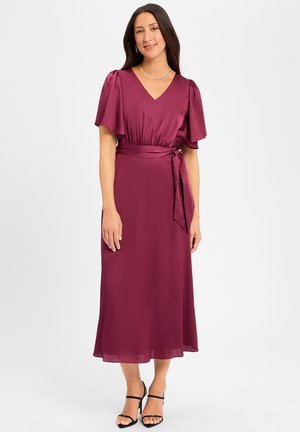 Burgunderrotes V-Ausschnitt-Kleid mit Puffärmeln, Taillenknoten und fließendem Rock. Glattes, seidiges Material mit mittellanger Länge bis zur Wade. Schwarze Riemchensandalen.