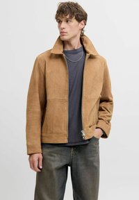 Tan ruskindsjakke med høj krave, lynlås og minimal syninger; båret over en mørkegrå skjorte og parret med afslappede jeans.