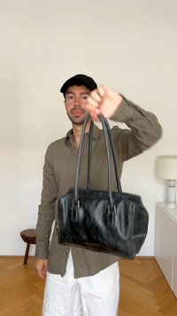 Schwarze Ledertasche mit zwei Griffen, strukturiertem Design, glänzender Oberfläche und auffälligen Nähdetails, gehalten von einer Person vor einem neutralen Hintergrund.