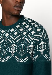 Sweter z dzianiny w kolorze teal, z białymi geometrycznymi wzorami, prążkowanym okrągłym dekoltem i teksturowanym materiałem, podkreślający zimowy design.