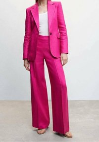 Różowy garnitur skrojony na miarę z usztywnianą marynarką i spodniami o szerokich nogawkach, wykonany z gładkiej tkaniny. Pod spodem biały top, noszony z płaskimi sandałami.