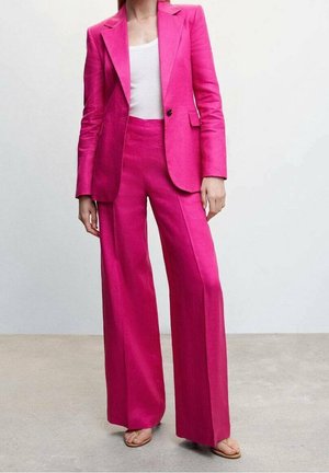 Vaaleanpunainen räätälöity puku, jossa on rakenteellinen bleiseri ja leveät housut, jotka on valmistettu sileästä kankaasta. Valkoinen toppi alla, yhdistettynä litteisiin sandaaleihin.