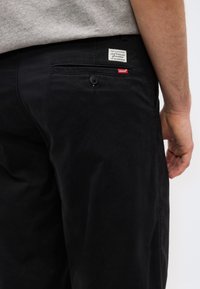 Άτομο που φοράει μαύρο παντελόνι Levi's με κουμπωμένη πίσω τσέπη και ορατή ετικέτα Levi's, σε συνδυασμό με γκρι πουκάμισο.