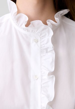 Primer plano de una blusa blanca con cuello alto con volantes y una volante vertical a lo largo de la tapeta de botones en la parte delantera.