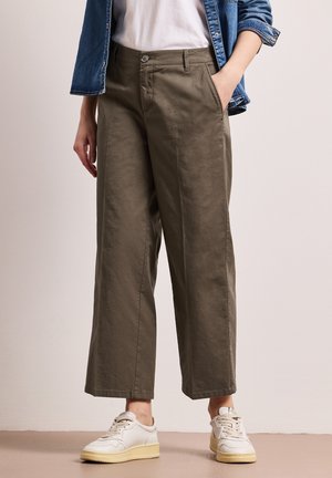 Person iført olivengrønne bukser med vide ben, hvide sneakers, hvid skjorte og blå denimjakke, med den ene hånd i lommen.