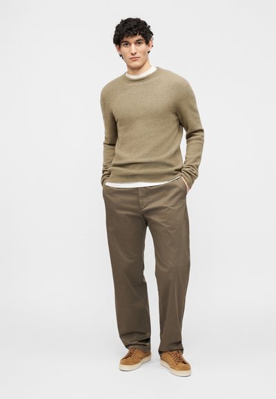 Άνδρας που στέκεται με τα χέρια στις τσέπες, φοράει πουλόβερ ελιάς, ανοιχτό καφέ παντελόνι, καφέ σουέντ αθλητικά παπούτσια και άσπρο εσώρουχο.