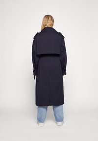 Cappotto trench blu navy con vita cinturata, spalline sulle spalle e design con spacco posteriore, abbinato a jeans azzurri chiari e sneakers bianche.