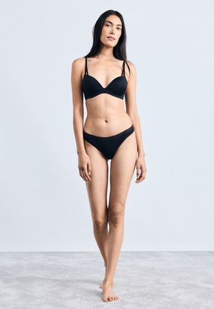 Zwarte beha en bijpassende bikini-achtige slip gemaakt van gladde stof. De beha heeft dunne bandjes en een diepe halslijn.