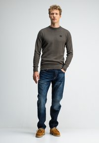 Junger Mann mit kurzen, lockigen Haaren steht in einem strukturierten braunen Pullover, blauen Jeans und hellen Wildlederschuhen vor einem schlichten weißen Hintergrund.