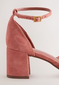 Sandale à talon bloc en daim rose avec bride de cheville et boucle dorée, présentant un talon fermé et un design à bout ouvert.