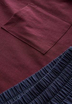 Burgunderfarvet stof har en lomme med skjulte syninger; nedenunder tilføjer en blød navy elastisk flæse tekstur og kontrast.