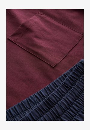 Burgunderfarvet stof har en lomme med skjulte syninger; nedenunder tilføjer en blød navy elastisk flæse tekstur og kontrast.