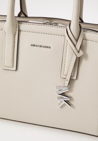 Lys beige lærhåndtaske med strukturet form, som har et sølv logo, et dekorativt mærke og kontrasterende syninger.