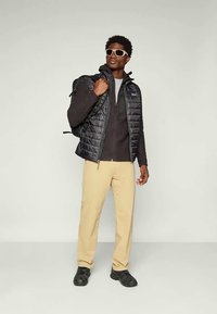 Man som bär solglasögon, svart pufferväst, mörk jacka, beiga byxor, svarta skor och bär en svart ryggsäck över ena axeln.