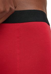 Shorts rouges ajustés avec une texture lisse et une large ceinture élastique noire. Le tissu semble extensible, idéal pour les vêtements de sport.