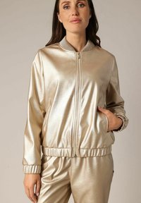 Veste bomber métallisée dorée avec fermeture éclair à l'avant, col côtelé, poignets élastiques et poches latérales ; fabriquée dans un matériau lisse et brillant.