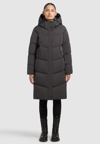 Sieviete, valkājot garu melnu ziemas jaku ar kapuci, melnas bikses un melnus zābakus, stāv pret vienkāršu gaišu fonu.