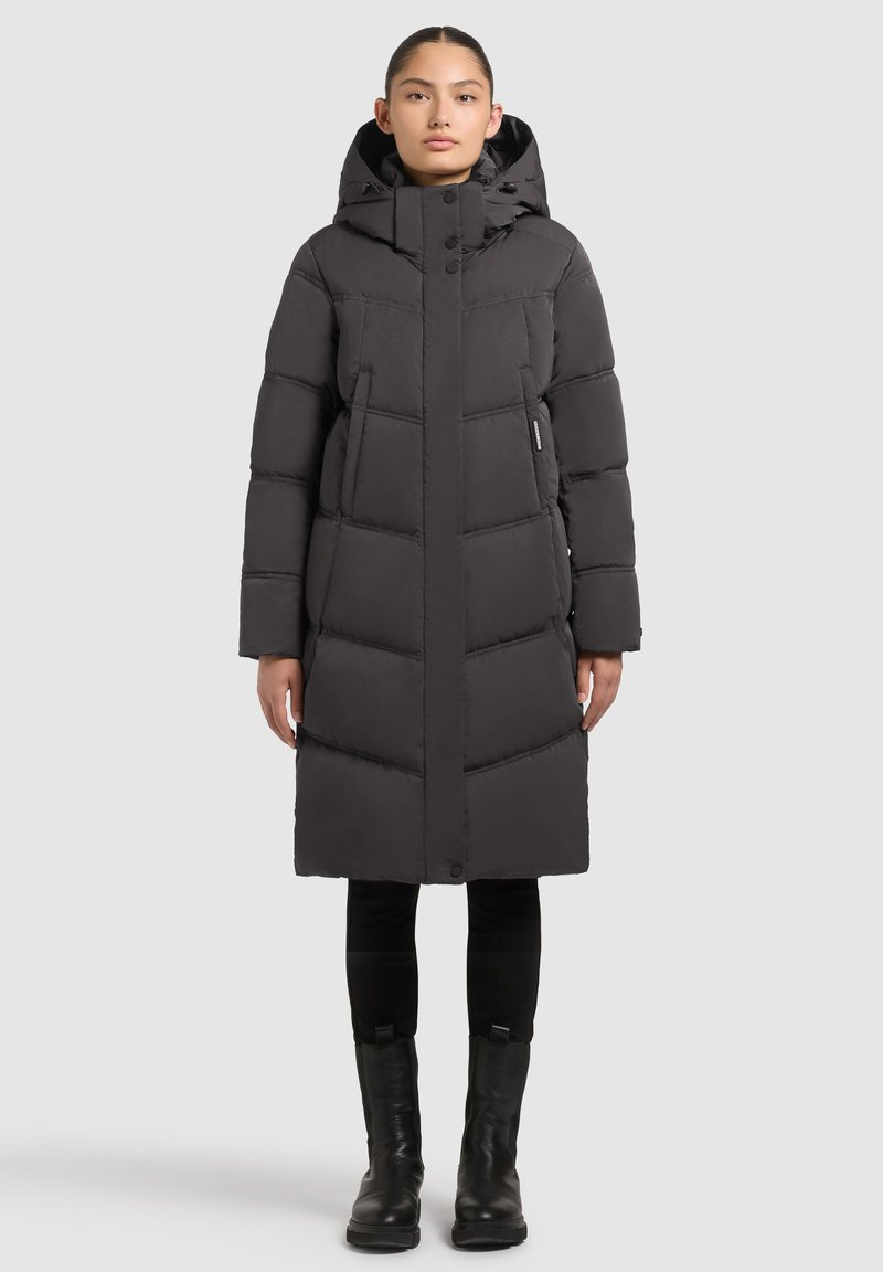 Sieviete, valkājot garu melnu ziemas jaku ar kapuci, melnas bikses un melnus zābakus, stāv pret vienkāršu gaišu fonu.