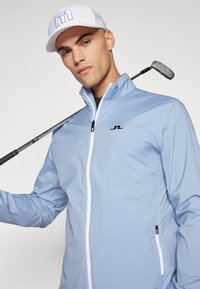Svijetloplava jakna na zip s visokim ovratnikom, kontrastnim bijelim zatvaračem i crnim logotipom. Model drži golf palicu i nosi bijelu kapu.