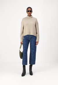 Beige hochgeschnittener, verkürzter Pullover mit hohem Rippkragen, kombiniert mit blauen, blumenmusternden weit geschnittenen Jeans und schwarzen Stiefeletten mit Absatz. Grünes Handtaschen-Accent.