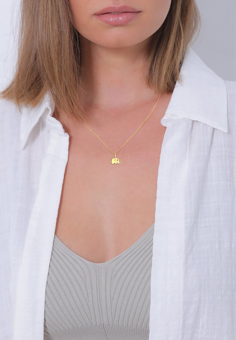Gouden ketting met een klein olifantenhangertje, gedragen over een lichtgrijze geribbelde top, onder een witte overhemd.