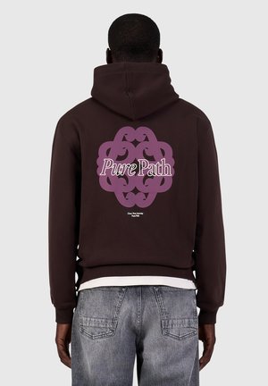 Man draagt een donkerbruine hoodie met een paarse decoratieve print en de tekst "Pure Path" op de achterkant, gecombineerd met vervaagde blauwe spijkerbroek.