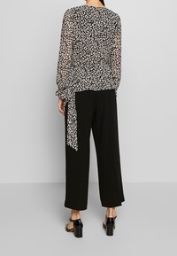 Top noir transparent avec imprimé léopard blanc, manches longues avec poignets élastiques, noué à la taille, associé à un pantalon noir à jambes larges et des chaussures à talons blocs.