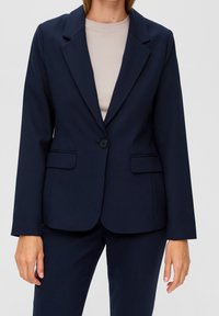 Marinblå, figursydd kavaj i glatt tyg, med enkel knappstängning, slagkrage och två framfickor. Bärs över en ljusbeige topp.