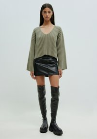 Olivengrüner, verkürzter Rippstrickpullover mit weiten Ärmeln, kombiniert mit einem schwarzen Leder-Minirock und knielangen schwarzen Stiefeln mit dickem Profil.