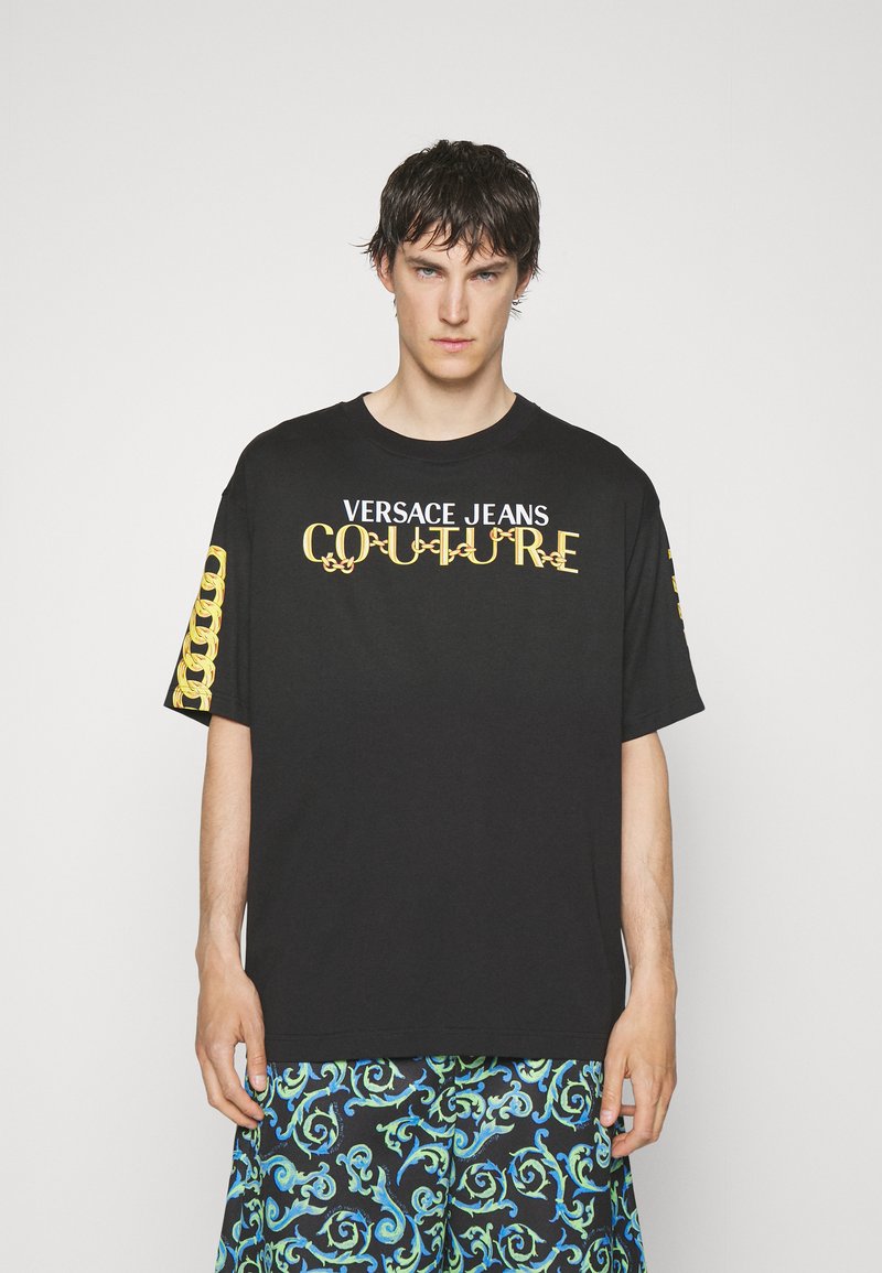 Schwarzes Baumwoll-T-Shirt mit dem Aufdruck "VERSACE JEANS COUTURE" in Gold. Verfügt über ein auffälliges dekoratives Muster auf den Ärmeln, einen lockeren Schnitt und einen Rundhalsausschnitt.