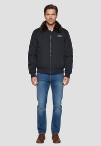 Giacca bomber blu navy con colletto in pelliccia sintetica, zip frontale, polsini e orlo a coste. Abbinata a jeans blu e scarpe marroni con lacci.