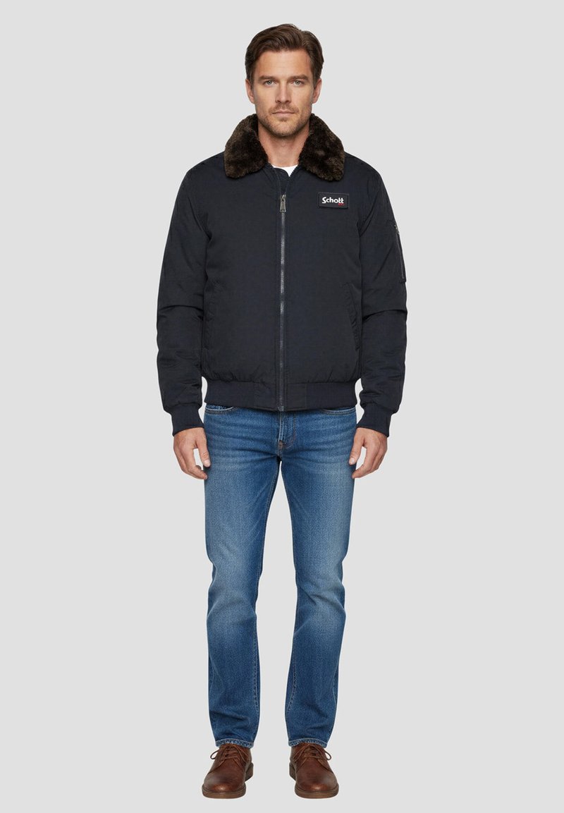 Giacca bomber blu navy con colletto in pelliccia sintetica, zip frontale, polsini e orlo a coste. Abbinata a jeans blu e scarpe marroni con lacci.