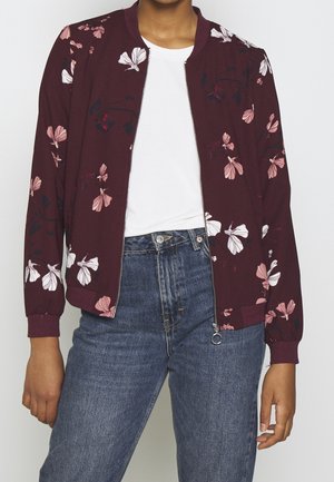 Femme portant une veste bomber bordeaux à motifs floraux avec fermeture éclair, un t-shirt blanc et un jean denim bleu, les mains détendues le long du corps.