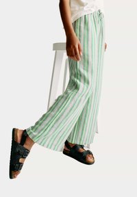 Gestreepte, lichte stoffen broek in groen en wit met roze accenten; losse pasvorm en elastische tailleband. Zwarte sandalen met twee gespen.