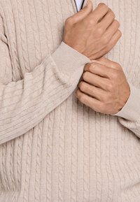 Maglione lavorato a maglia beige con motivo a trecce, polsini a coste e collo con zip. Il tessuto appare morbido con una leggera texture.