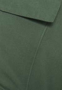Tecido verde com uma textura suave, apresentando um colarinho dobrado e detalhes de costura visíveis. O material parece durável e leve.